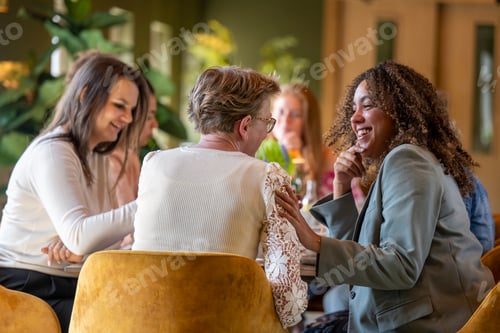 Preview: Group of women enjoying a cheerful conversation on yellow velvet chairs with drinks in hand.