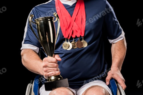 Preview: cropped view of paralympic in wheelchair with champion goblet and gold medals isolated on black