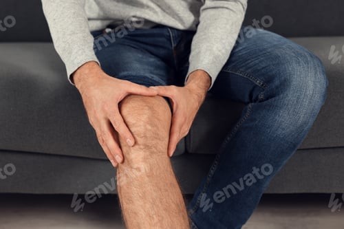 Preview: Man suffering from leg pain on sofa indoors, closeup