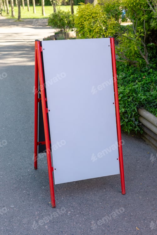 Preview: A blank outdoor sign stands on a pathway in a park filled with greenery.