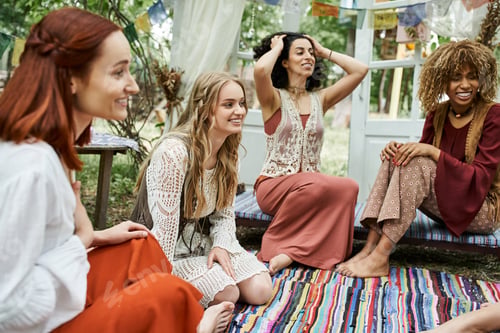 Preview: cheerful and multiethnic women in boho styled clothes talking outdoors in retreat center