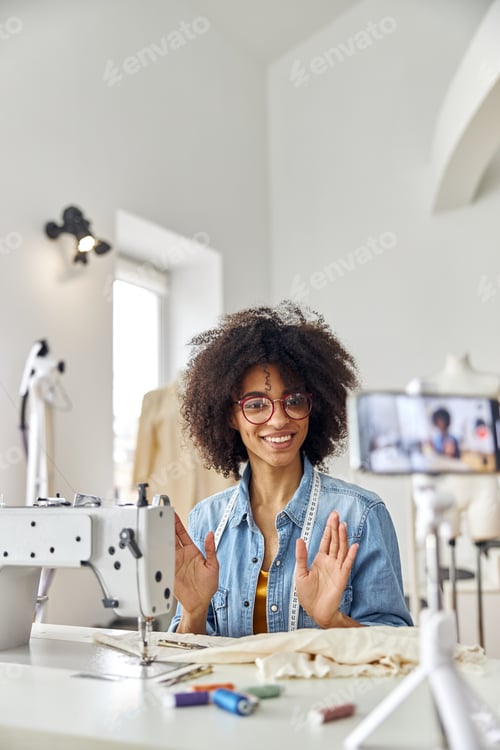 Preview: African-American woman dressmaker looks at phone camera at workplace with sewing machine in studio