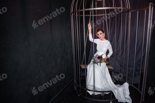 Preview: Fashion bride sits on swing in an iron cage, peeking out from behind the bars. Fictitious marriage