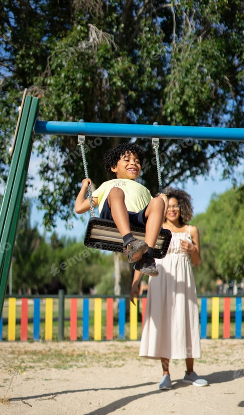 Preview: Latino boy with afro hair swinging on a playground swing with his mother on a sunny day