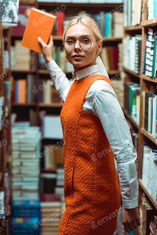 Preview: blonde and beautiful woman in glasses and dress holding book and looking at camera in library