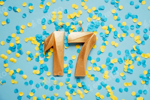 Preview: Number 17 seventeen golden celebration birthday candle on yellow and blue confetti Background.