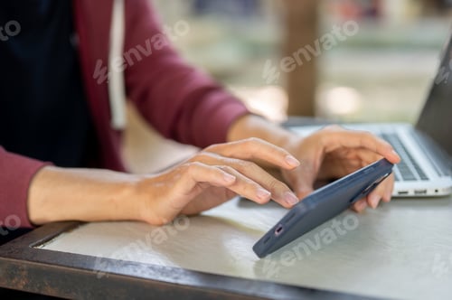 Preview: Close up of hand holding and touching on smartphone while sitting with a laptop at concrete table.