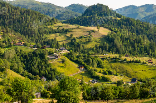 Preview: Lush green valley with rural houses surrounded by forested mountains under clear blue sky.