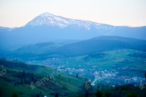 Preview: Scene of valley village surrounded by verdant hills, with majestic, snow-capped mountain.