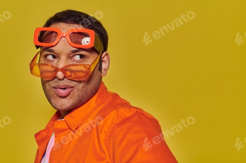 Preview: fashionable indian man trying on different trendy sunglasses and looking away on yellow backdrop