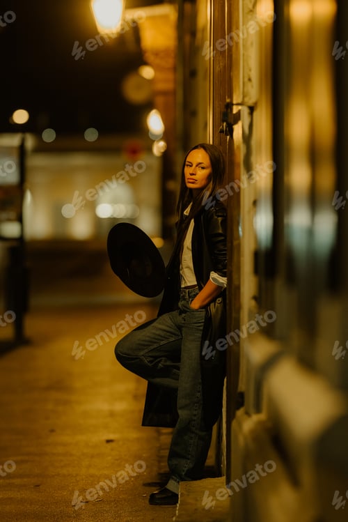Preview: Stylish woman leaning against wall at night holding hat