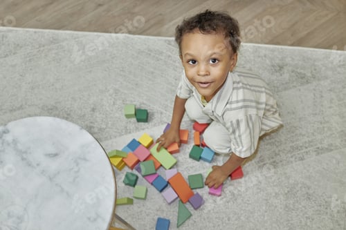 Preview: Little Black Boy Smiling at Camera Playing with Building Bricks while Sitting on Floor at Home