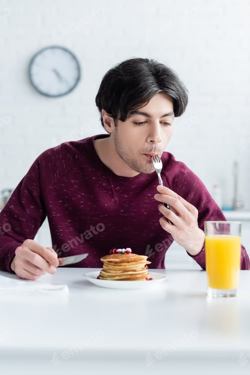 Preview: young brunette man eating tasty pancakes near glass of fresh orange juice