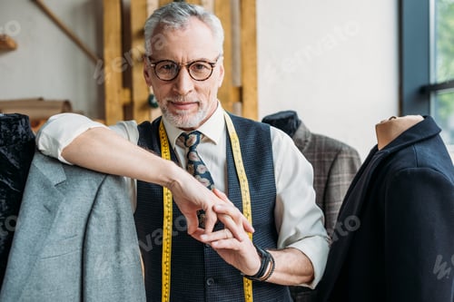 Preview: smiling mature tailor with measuring tape leaning on mannequin with jacket at sewing workshop and