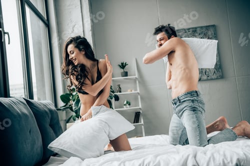 Preview: cheerful brunette girl in lace underwear having pillow fight with happy muscular boyfriend on bed