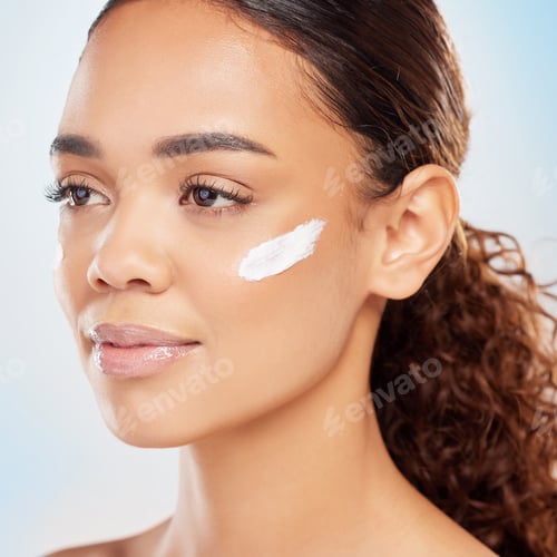 Preview: Shot of a young woman applying cream to her face against a blue background