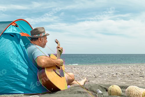 Preview: Middle aged bearded man playing guitar and singing while camping picnic on beach in summer vacation.