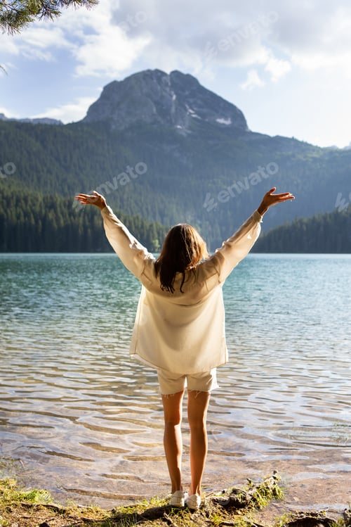Preview: The girl stands with her back near a mountain lake in the background. Positive young woman traveling