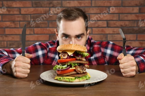 Preview: Young hungry man with cutlery eating huge burger at table