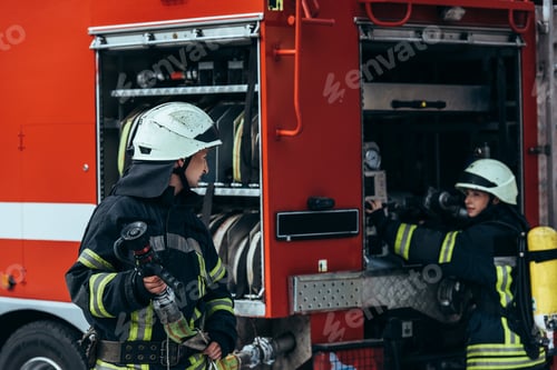 Preview: male firefighter in uniform holding water hose while colleague checking equipment in truck on street