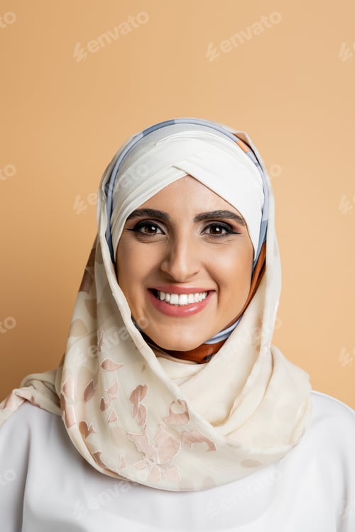 Preview: portrait of charming muslim woman with makeup, in silk headscarf smiling at camera on beige