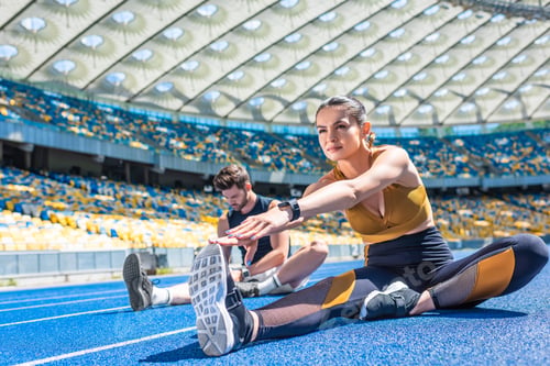 Preview: young male and female joggers sitting on running track and stretching at sports stadium