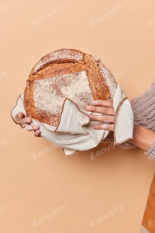 Preview: Vertical shot of unrecognizable woman holds round loaf of bread on towel isolated over beige backgro