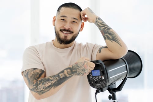 Preview: Smiling asian photographer setting up his lighting looking at camera in photo studio
