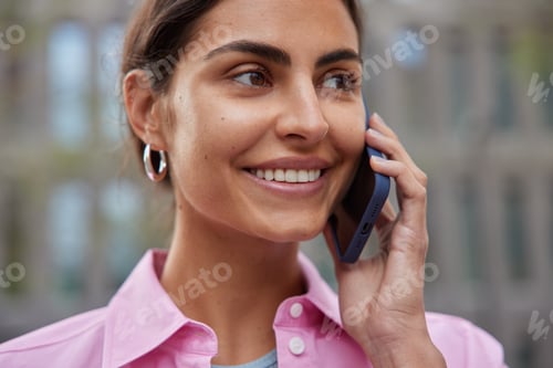 Preview: Close up shot of happy positive beautiful woman smiles gently keeps modern smartphone near ear has p