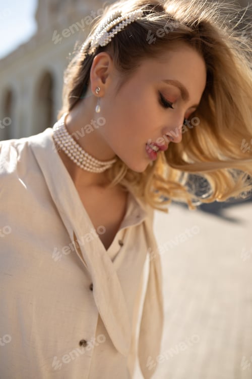 Preview: Charming curly blonde woman in pearl jewelry and beige blouse looks down, walk outside. Young girl