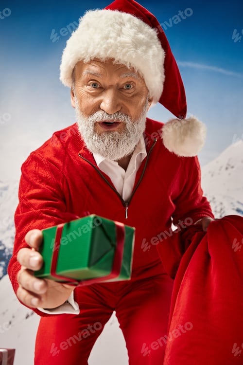 Preview: white bearded Santa Claus lending present with red ribbon and looking at camera, winter concept