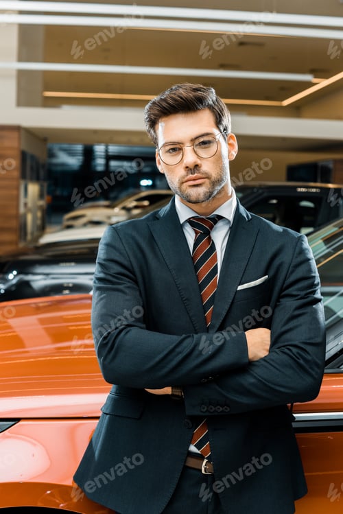 Preview: confident businessman in eyeglasses posing with crossed arms near automobile