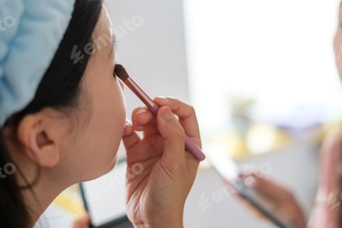Preview: Asian woman doing makeup with brush in front of bathroom mirror.