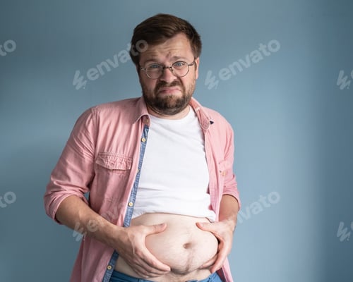 Preview: Unhappy, dissatisfied overweight man, lifted t-shirt and holds excess belly fat with his hands