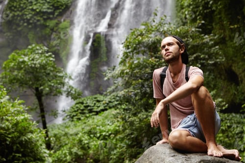 Preview: People, nature and adventure. Young hipster with backpack sitting on big rock by waterfall on backgr