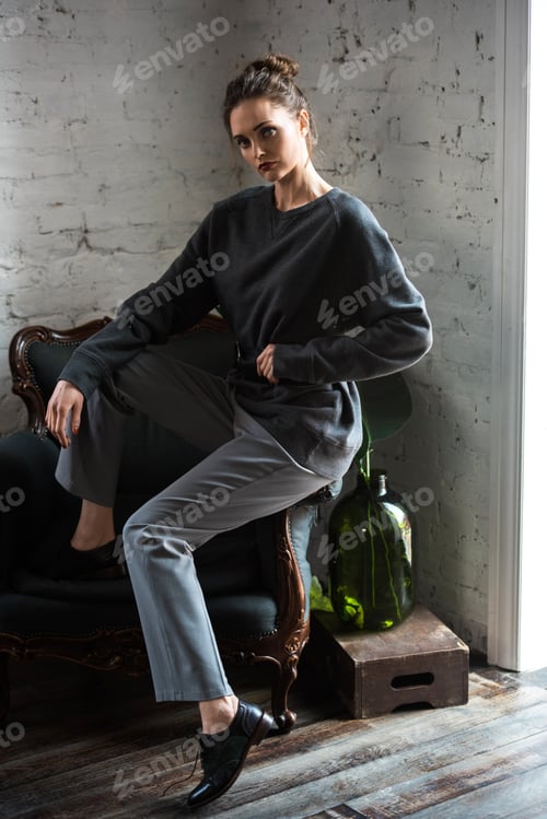 Preview: full length view of attractive brunette girl in trendy outfit sitting on armchair and looking at
