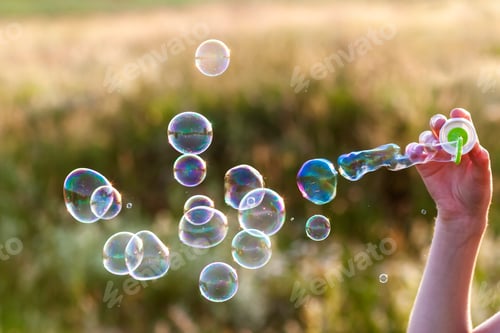 Preview: Womans hand with blowing colorful soap bubbles at sunset.