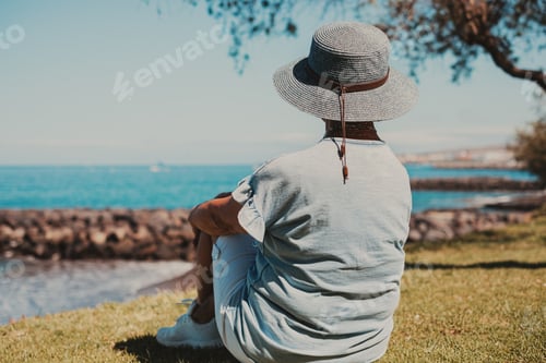 Preview: Back view of mature caucasian woman sitting in the meadow close the beach looking horizon over sea