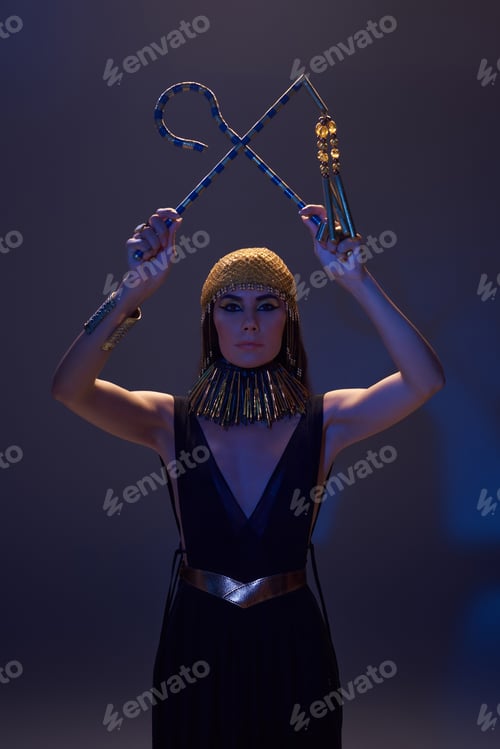 Preview: Woman with egyptian makeup and look holding crook and flail on brown background with blue light