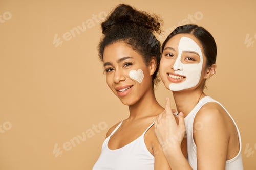 Preview: Two young female friends having fun, smiling at camera while posing with facial masks on isolated