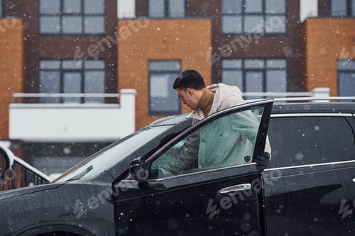 Preview: Young handsome man outdoors near his modern black car at winter time