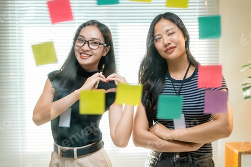 Preview: Asian businesswomen making heart shape with hands near sticky notes on glass wall in office