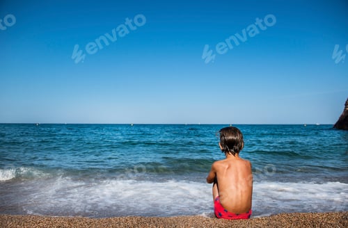 Preview: Rear view of boy sitting on beach looking out at sea, Begur, Catalonia, Spain