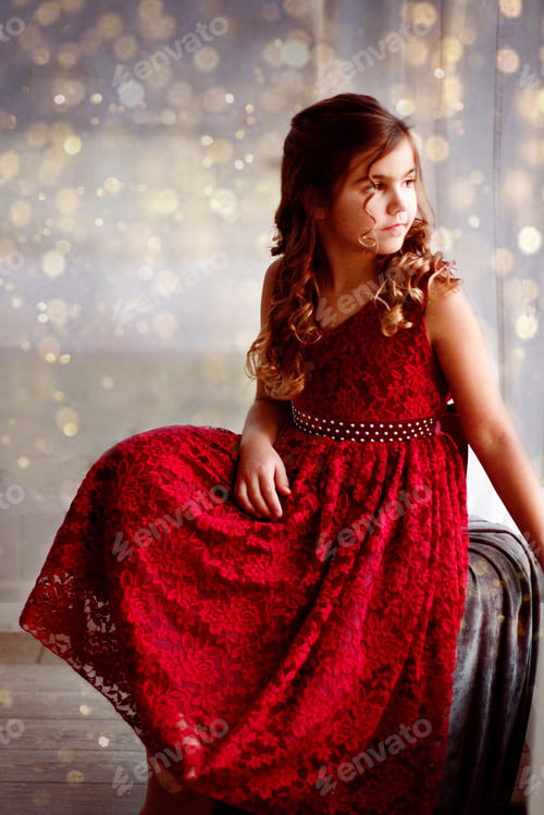 Preview: Girl in Christmas Dress looking out the window with bokeh lights around her.