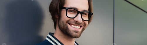 Preview: pleased man in trendy eyeglasses looking at camera near grey wall, banner