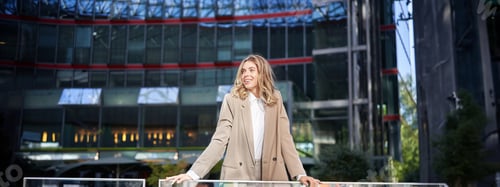 Preview: Young beautiful businesswoman standing in beige suit on sunny street near office buildings