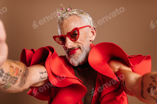 Preview: Confident mature gay man in beautiful red dress making self portrait against brown background
