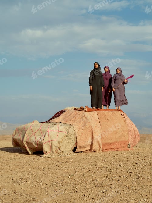 Preview: Three women dressed in traditional attire stand beside a covered car in a desert landscape.