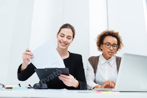 Preview: Two happy businesswomen using clipboard and laptop together