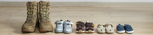 Preview: Old worn military boots and lot of baby shoes on wooden floor. Military father and family concept.